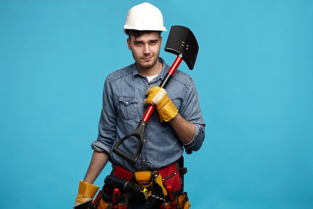 Foto van jonge graafmachine die handschoenen, witte helm en hulpmiddelriem dragende schop draagt