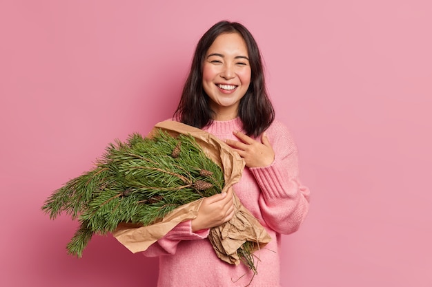 Gratis foto foto van knappe brunette vrouw glimlacht in het algemeen voelt tevreden houdt fir boomtakken heeft feeststemming gonna make kerst samenstelling draagt winter trui
