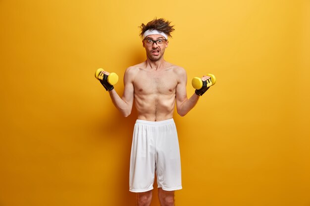 Foto van magere man gaat sporten, bouwt thuis spieren op, heeft een effectief trainingscomplex met halters, draagt een witte korte broek, poseert met blote torso tegen gele muur. Gezondheidszorg concept
