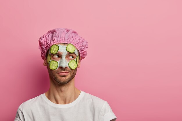 Foto van mannelijk model opzij geconcentreerd, heeft schoonheidsprocedures, past kleimasker en komkommers toe op de teint