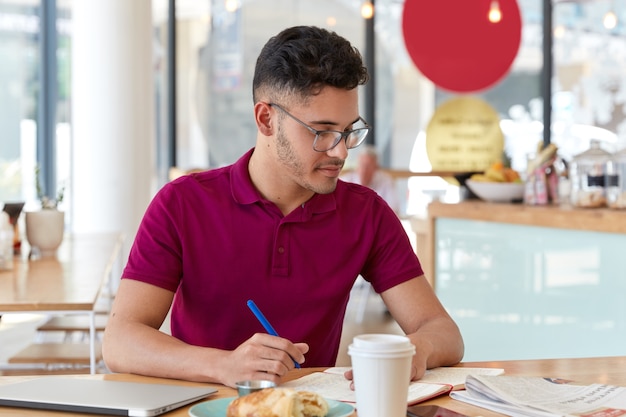 Foto van mannelijke student van gemengd ras schrijft noodzakelijke informatie in Kladblok uit de dagelijkse krant, maakt soortgelijk artikel, zit binnen tegen café-interieur, drinkt afhaalmaaltijden koffie, leert binnen