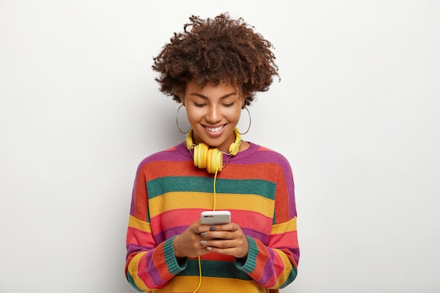 Foto van mooie Afro-Amerikaanse vrouw geconcentreerd in smartphoneapparaat, geniet van online chatten, downloadt muziek naar afspeellijst om te luisteren in een koptelefoon, heeft donker krullend haar, draagt casual outfit