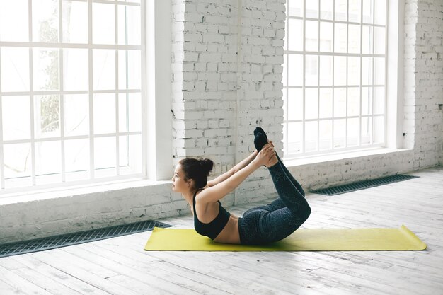 Foto van mooie fitte jonge gevorderde vrouwelijke yogi in stijlvolle sportkleding die 's ochtends Ashtanga-yoga beoefent in een ruime lichte hal, booghouding of dhanurasana doet. Mensen en een gezonde levensstijl