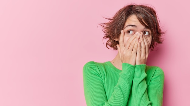 Foto van mooie jonge Europese vrouw met kort haar bedekt gezicht met handen probeert te verbergen voor iets draagt groene trui geïsoleerd over roze achtergrond lege kopie ruimte voor uw promotie