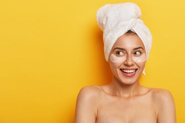 Foto van mooie natuurlijke vrouw met make-up, kijkt opzij met zachte glimlach, is van toepassing op plekken onder de ogen voor het droge huidtype, draagt een handdoek