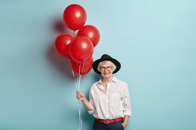 Foto van mooie oude vrouw heeft plezier op feestje met mensen van dezelfde leeftijd, houdt bos luchtballonnen vast, draagt stijlvolle hoed, wit overhemd en zwarte broek, vormt over blauwe muur, viert verjaardag