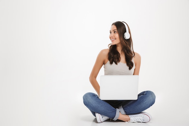 Foto van mooie vrouw die aan muziek luistert of het gebruiken van hoofdtelefoons en laptop babbelt terwijl het zitten met benen op de vloer, over witte muur worden gekruist
