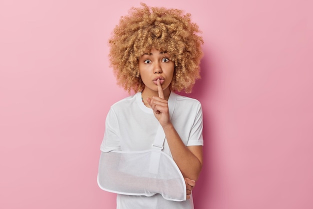 Foto van mysterieuze vrouw maakt stiltegebaar houdt wijsvinger over lippen vertelt geheim heeft gebroken arm in spalk gekleed in casual wit t-shirt geïsoleerd over roze achtergrond gewond vrouwelijk model