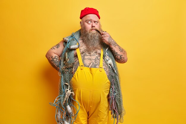 Foto van nadenkende mollige zeeman poseert met visnet rookt pijp trekt wenkbrauwen op met peinzende uitdrukking gekleed in overall getatoeëerd lichaam