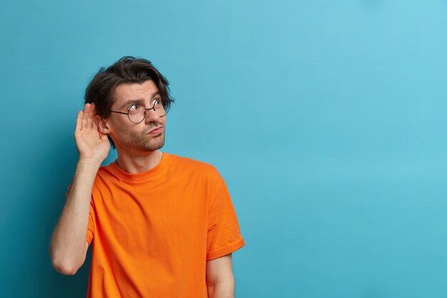 Foto van nieuwsgierige man houdt de hand in de buurt van het oor en luistert naar privé-informatie, probeert roddelen af te luisteren, heeft een geïntrigeerde uitdrukking, draagt een ronde bril en een oranje t-shirt, kopie ruimte op blauwe muur