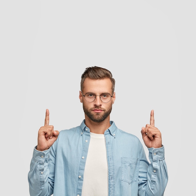 Foto van ongeschoren hipster gekleed in casual shirt, wijst met beide wijsvingers naar boven.