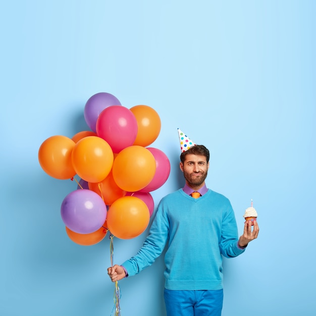 Foto van ongeschoren man met verjaardagshoed en ballonnen poseren in blauwe trui