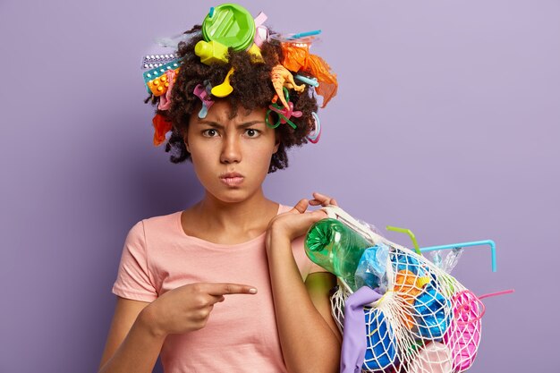 Foto van ontevreden Afro-Amerikaanse vrouw die boos is op misbruik van plastic, wijst naar zak met verzameld afval, heeft afval in het hoofd, geïsoleerd op een paarse muur. Niet recyclebaar vervuilingsconcept