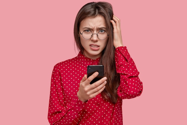 Foto van ontevreden donkerharige vrouw kijkt verbaasd, houdt mobiele telefoon vast, denkt na over de inhoud van het bericht, gekleed in elegante kleding