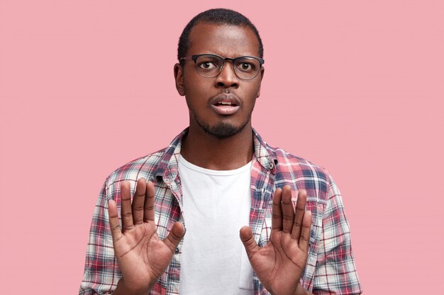 Foto van ontevreden man met donkere huid en ontevreden uitdrukking, maakt weigeringsgebaar, vraagt om ermee te stoppen. Knap African American man in bril gebaren