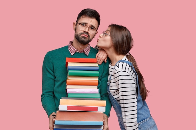 Foto van ontevreden ongeschoren man draagt schoolboeken, ontvangt kus, draagt een bril, geïsoleerd over roze ruimte