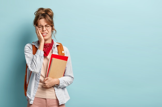 Foto van ontevreden stressvol moe schoolmeisje draagt spiraalvormige blocnote en boek, draagt een ronde bril