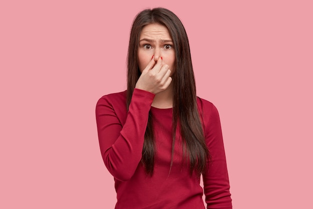 Foto van ontevreden vrouw sluit neus met stank, voelt vreselijke geur van afval, draagt rode kleren