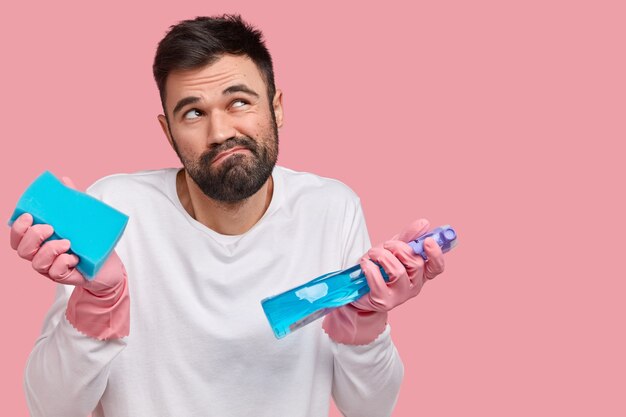 Foto van onwetende ongeschoren man heeft een onzekere blik, fronst zijn wenkbrauwen, is naar boven gericht, draagt een spons en een schoonmaakproduct