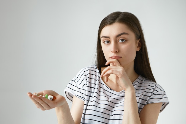 Foto van onzekere jonge brunette vrouw die een mondvol kleurrijke pillen vasthoudt, een bedachtzame twijfelachtige uitdrukking heeft, de kin aanraakt, eraan denkt medicatie te nemen of niet terwijl ze aan verkoudheid lijdt