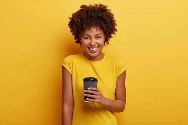 Foto van opgetogen Afro-Amerikaanse vrouw houdt afhaalkoffie, geniet van aromatische drank, heeft brede glimlach, witte tanden, draagt casual geel t-shirt