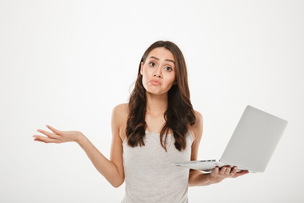 Foto van overstuur vrouw met lang bruin haar handen overgeven en verwarring uitdrukken terwijl zilveren personal computer, geïsoleerd over witte muur