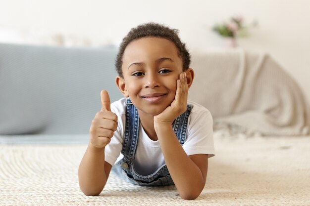 Foto van positief vriendelijk ogende acht-jarige Afro-Amerikaanse jongen liggend op de vloer thuis