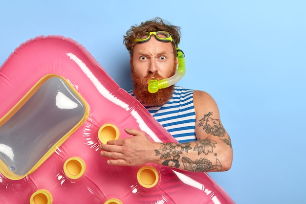 Gratis foto foto van roodharige gember man draagt zwembril, snorkelmasker, gaat duiken en zwemmen in zee, houdt roze opgeblazen matras vast, ziet er serieus uit