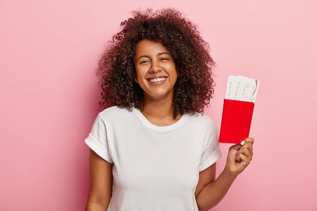 Foto van schattige donkere vrouw houdt kaartjes en paspoort, verheugt zich op zomervakantie en reis, blij dat haar droom eindelijk uitkwam, gekleed in een wit t-shirt, wacht op het vliegtuig. reizend concept