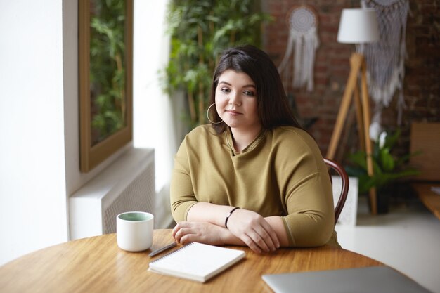 Foto van stijlvolle overgewicht jonge vrouwelijke ontwerper zittend op een naaiatelier en koffie drinken, nadenken over concept van nieuw project, schetsen in dagboek, nadenkend doordachte blik hebben