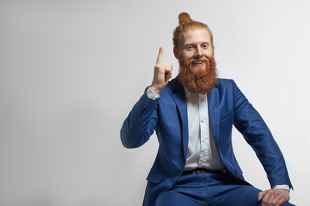 Foto van stijlvolle zelfverzekerde jonge mannelijke CEO met haarknoop en dikke gemberbaard glimlachend gelukkig naar boven gericht, zittend tegen lege studio muur achtergrond met kopie ruimte voor uw informatie