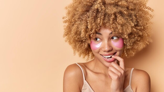 Foto van tevreden dromerige vrouw bijt vinger kijkt weg met peinzende uitdrukking past roze hydrogelpleisters toe voor huidbehandeling geïsoleerd op beige achtergrond mockupruimte voor uw promotie