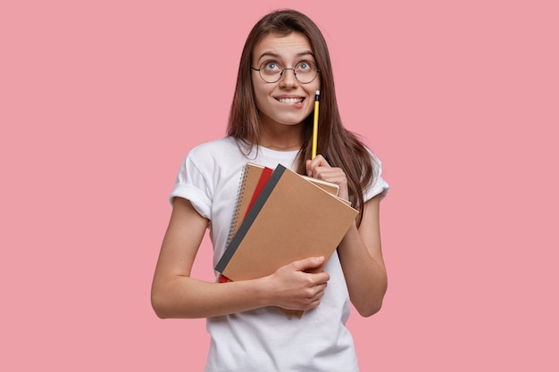 Foto van tevreden europese jonge dame kijkt gelukkig naar boven, houdt potlood, schoolboeken vast, heeft dromerige uitdrukking, schrijft notities