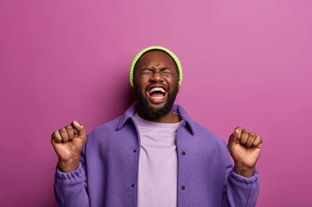 Foto van tevreden gelukkig hipster man met opgeheven vuisten, heeft plezier op feestje, voelt zich overmatig, vrolijk zijn, loenst van het lachen, voelt als kampioen