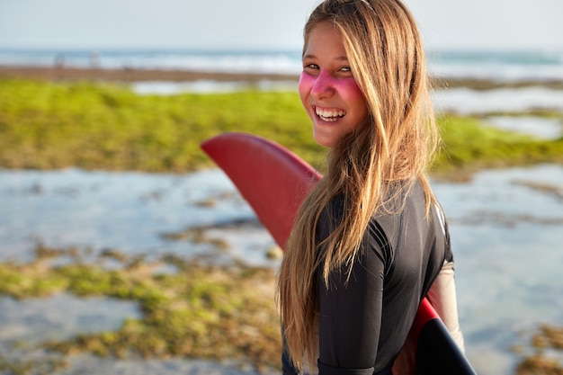 Foto van tevreden langharige vrouw houdt surfplank onder de hand, ziet er positief uit