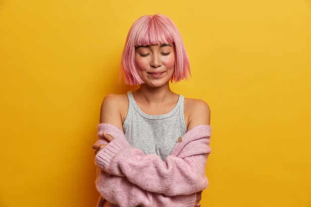 Foto van tevreden vrouw met bob kapsel, roze haar sluit ogen van plezier, draagt zachte warme trui die van de schouders is gevallen