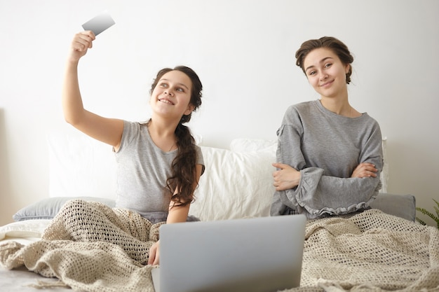 Foto van tienermeisje en haar moeder of oudere zus zittend op bed voor geopende laptop, online winkelen, eten bestellen of nieuwe kleren kopen
