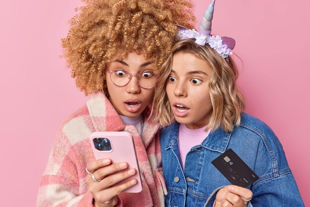 Foto van twee geschokte mooie vrouwen staren naar smartphone, creditcard vasthouden, verwacht geen bonus en cashback online betalen, gekleed in modieuze kleding geïsoleerd op roze achtergrond