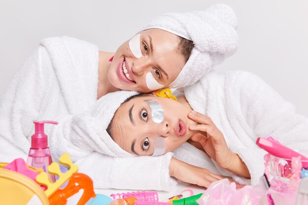 Foto van twee jonge vrouwen van gemengd ras die met hun hoofd poseren bij een tafel met cosmetische producten die pleisters onder de ogen aanbrengen