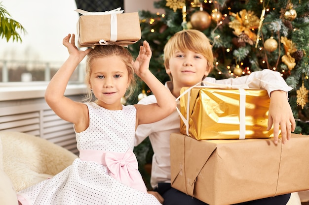 Foto van twee schattige Europese kinderen broers en zussen poseren bij de kerstboom. Knappe tiener die nieuwjaarsgeschenken uitpakt samen met zijn schattige zusje naast hem met een doos op haar hoofd