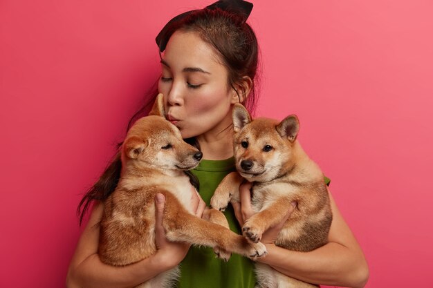 Foto van verantwoordelijke vrouwelijke gastheer kust schattige puppy's, draagt naar trimsalon of dierenarts, bezoekt hondensalon, heeft vriendschappelijke relatie, poseert tegen roze achtergrond