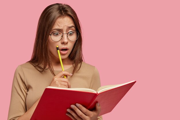 Foto van verbijsterde aantrekkelijke vrouw leest schokkende informatie in leerboek, houdt potlood vast, houdt mond wijd open