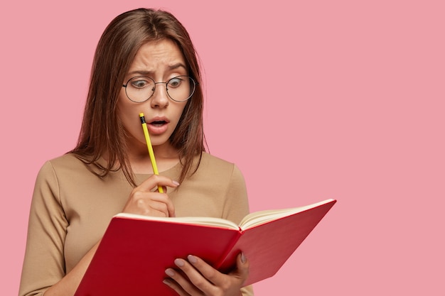 Foto van verbijsterde aantrekkelijke vrouw leest schokkende informatie in leerboek, houdt potlood vast, houdt mond wijd open