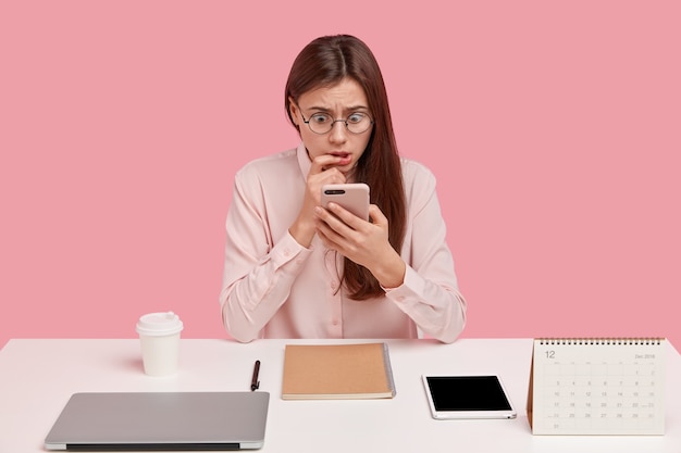 Foto van verbijsterde Europese vrouw bladert op internet op moderne mobiele telefoon, heeft bang expressie, poses op de werkruimte, gekleed in een formeel overhemd