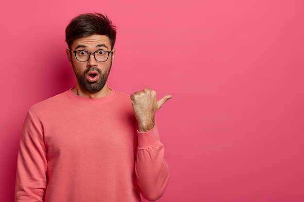 Foto van verbijsterde jonge, bebaarde man geeft aan de rechterkant aan, toont iets verbazingwekkends, hijgt naar adem van verwondering, draagt een bril en een losse trui, poseert tegen een roze muur. Promotie concept