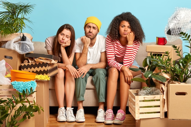 Gratis foto foto van vermoeide drie verschillende studenten verhuizen naar een nieuwe flat, veranderen van woonplaats, hebben slaperige ontevreden uitdrukkingen, vermoeidheid na het uitpakken van bezittingen, omringd door items in kartonnen containers