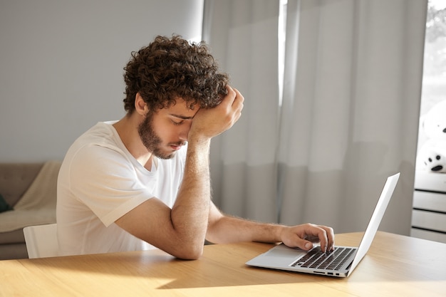 Foto van vermoeide jonge mannelijke vertaler met dikke baard en golvend haar hoofd aanraken en ogen sluiten, zich absoluut uitgeput voelen, toetsen op laptop, technische documentatie vertalen