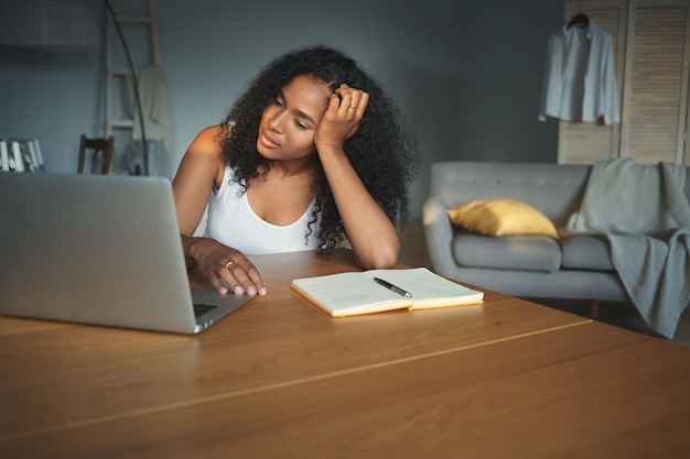 Foto van vermoeide mooie jonge Afrikaanse vrouwelijke student zittend aan een bureau met open elektronisch apparaat, zich uitgeput voelen tijdens het prearing om te testen. Mensen, technologie, opleiding, baan en beroep