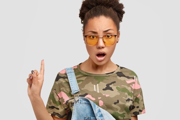 Foto van verontwaardigde donkere vrouw met ontevreden uitdrukking, opent mond en toont iets naar boven, nonchalant gekleed, poseert over een witte muur.