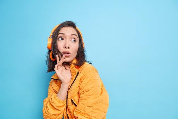 Foto van verrast gefascineerd Aziatisch tienermeisje gefocust met bang geschokte uitdrukking boven gekleed in oranje jasje luistert muziek uit afspeellijst via koptelefoon geïsoleerd over blauwe muur